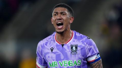 Liam Palmer looks ahead with his motuh wide open shouting. he wears a purple short sleeve football shirt and captains armband.