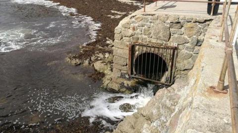 A pipe at a beach in Cornwall. The pipe is coming from beneath a platform overlooking the sea. The hole is covered by a metal grate and liquid is pouring out of the pipe and into the sea.