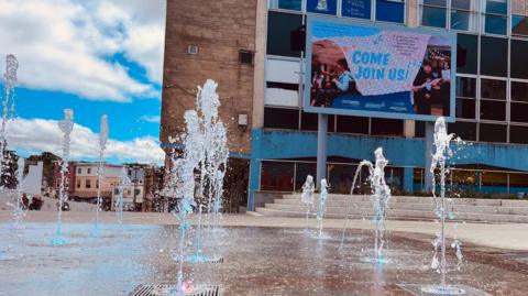Water jets shoot upwards between paving slabs in a wide open space. The ground is wet due to the jets and a large, run-down building is seen behind.