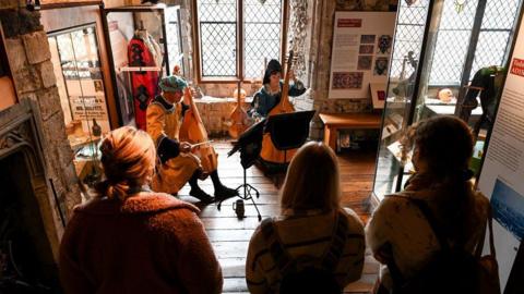 A group of three people watch two people sitting playing instruments while dressed in medieval clothing. They are all inside Westgate Museum.