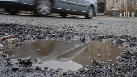 A generic image of a pothole in a road as a silver car passes nearby. The pothole has been filled with rainwater.