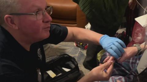 The picture shows a paramedic providing medical care to an elderly person in their own home. The paramedic is wearing protective gloves and has a medical kit open beside them on the floor, along with monitoring equipment. The setting looks like a living room, with everyday household furniture around them, suggesting the care is being delivered in the community rather than in hospital.