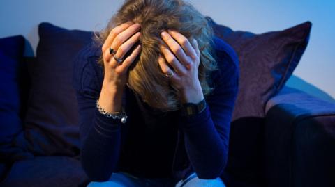A woman is sat with her head in her hands on a dark blue sofa. She has dark blonde hair and silver rings on her fingers.