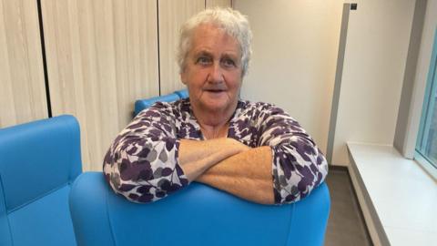 Ann has short white hair. She is wearing a floral blouse and leaning on a blue chair.