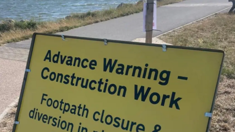 A yellow temporary roadworks sign which reads: 'Advance Warning - Construction Work. Footpath closure and diversion.' In the background is the riverside path and the water of Parkstone Bay in Poole Harbour.
