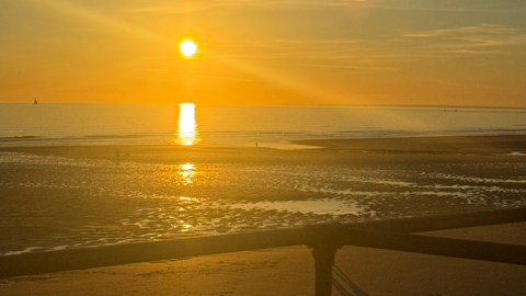The sun setting on a low tide at St Ouen's bay with the handrail in the foreground and a small boat to the left in the ocean. People walking along the beach.