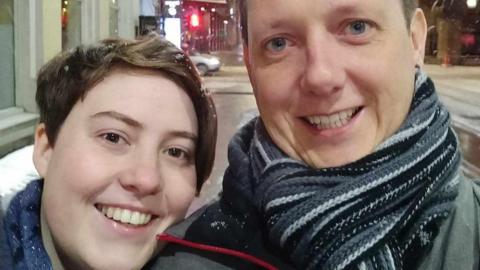 A man with light-blue eyes, blond hair, black, grey and white scarf, and a woman with short brown hair and brown eyes. Both are smiling. 