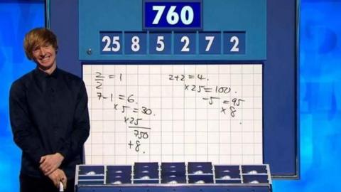 Dr Tom Crawford on the Countdown set. On the white board is some arithmetic, with the challenge to make the number 760 from 25, 8, 5, 2, 7 and 2. He has light brown hair brushed across his forehead and has a lip piercing and ear studs. He wears a long dark blue, long-sleeved shirt, buttoned to the top.