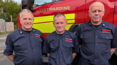 Three brothers stand in a row in front of a red fire engine that says "Leintwardine" in gold letters. They are all dressed in navy fire shirts with a red logo.