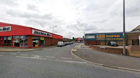 Google Street View of the junction of Ellis Way and Holles Street. There are a number of warehouse style shops on the corner