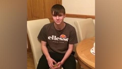 George Joseph Powell sitting on a cream chair, wearing a brown T-shirt and smiling into the camera 