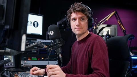 Greg James wears a burgundy jumper. He is sitting in front of a radio desk with headphones on. 