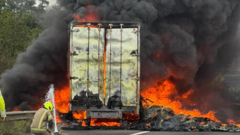 Firefighter points a hose of water at a burning lorry surrounded by thick black smoke and red flames