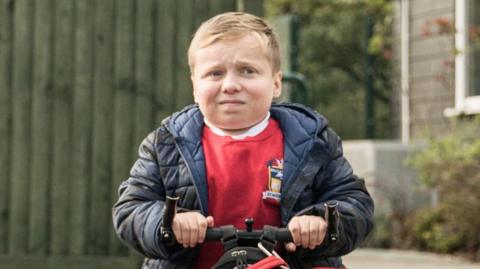 A boy, the character Ollie, grimaces - he wears a red school jumper and blue coat