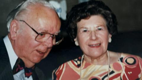 Una Crown and her husband, Ron. He is on the left and almost in profile as if in the middle of talking, wearing glasses with swept back white hair and wearing a dark jacket, white shirt and blue and red bow tie. She is on the right with dark short curling hair, wearing big stud earrings, a pink, red, beige and black patterned dress and a long string of pearls. She is about to smile.
