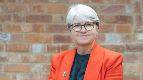 Anna Earnshaw with short white hair and brown-framed glasses wearing a dark top and red jacket with a small broach on the right lapel. She is looking at the camera and standing in front of a brick wall.