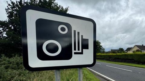 A black and white speed camera ahead sign mounted on a grass verge at the side of a single lane asphalt carriageway.