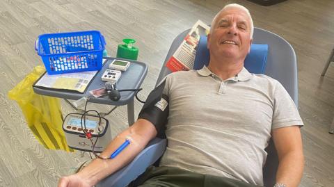 A man with short white hair smiles at the camera as he lies back in a medical chair with a needle in his arm connected to a tube carrying blood to a collection device. He is wearing a grey polo shirt and has a blood pressure monitor on his arm. A small table connected to the side of the chair has medical devices and a blue plastic basket on it. 