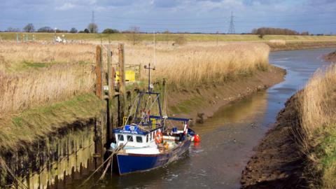 The mouth of the Vernatt’s Drain where it feeds into the River Welland near Spalding, The eastern bank of the Welland can be seen crossing the centre of the image and there is a blue and white boat in the foreground.