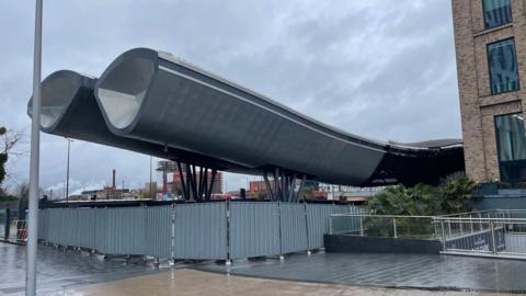 The distinctive curved roof of the 70 metre long aluminium clad bus station looms dramatically against a dark, foreboding looking sky. Dull grey metal fencing surrounds the now derelict building with no people, let alone any passengers in sight. 