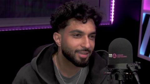 Tegi Pannu, a male singer, in an interview with BBC Asian Network. He is wearing a black hoody, grey tshirt with a silver necklace, while speaking into a BBC Asian Network branded microphone. Behind him is a wall with a purple LED light.