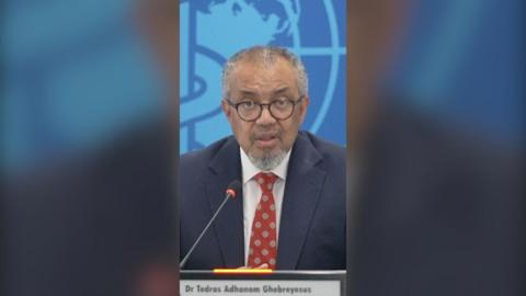 Tedros Ghebreyesus looks at the camera as he speaks at a press conference. He wears a navy blazer, white shirt and red tie.