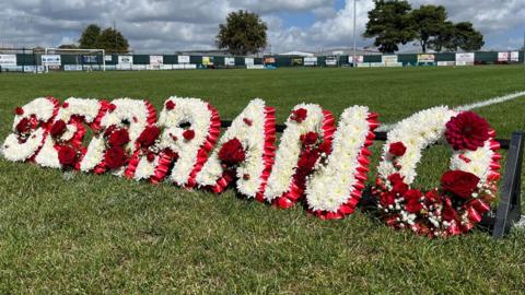 A floral tribute in red and white flowers spelling out Serrano. The letter are resting on a black metal frame on the green grass of Thetford Town Football Club's pitch. 