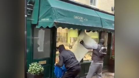 A jewellery shop window is being ripped off by a man while another is bent down with a blue bag