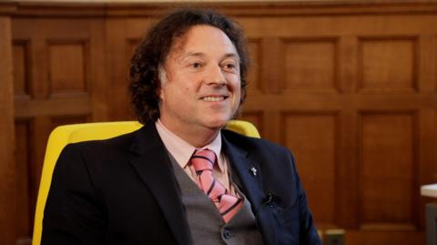 A man in a suit with a pink tie. He is sat in a chair in a wood-panelled room and is looking to the side of the frame.