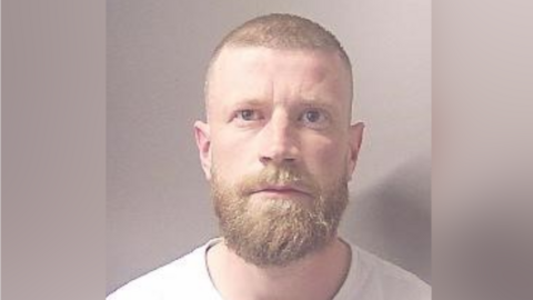 Hodson is in custody and stares straight at the camera. He is wearing a light top and has short hair and a large beard. He is in front of a plain light beige background.