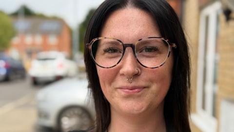 Natasha Saggs - a woman with nose piercings, large glasses and black hair smiles at the camera.