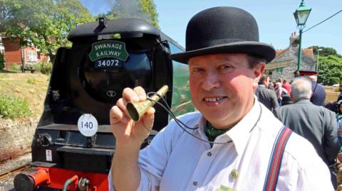 A smiling Peter Sills posing for the camera and holding up the brass horn, which is the size of a large cigar, between his thumb and fingers. He is wearing a white shirt and cravat and a black bowler hat. Behind him is a black and green steam locomotive with Swanage Railway 34072 written on the front.