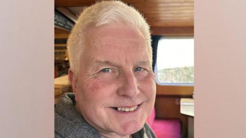 Martin Ball is seated and is smiling at the camera. He has short white hair and is wearing a grey hoodie