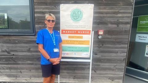 A nurse in a blue top is standing outside a wooden office by a sign that reads Market Nurse here today. No appointment needed - Free! Penbode Hut. To the far right of the photo there is an open door with "market nurse" written on it and a poster for Penbode.