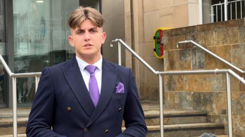 A man standing in a navy blazer and purple tie, on the steps of a council building built in light sandstone.