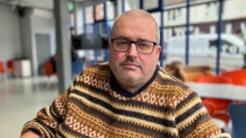A man with a shaved head in square glasses and a brown knitted jumper sitting in a cafe and looking at the camera with a neutral expression
