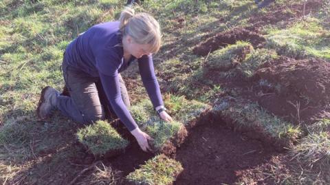 A woman knelt on the grass making a test pit. There is a square of turf that has been pulled apart in squares. She is looking down and is wearing a purple long-sleeved top.