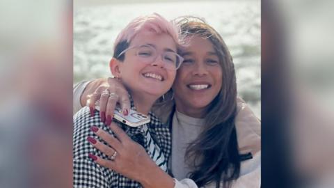 Too women smile at the camera, the one on the right has her arms wrapped around the other. The sea is behind them with the sun shining off it.