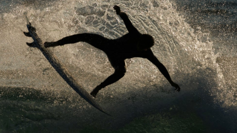 The picture shows a surfer performing an aerial manoeuvre on a wave. The surfer is silhouetted against the sunlight with the spray of water that arcs behind them. The surfboard is angled upward, and the surfer’s body is extended with arms outstretched. The background is the ocean with shimmering light on the surface.