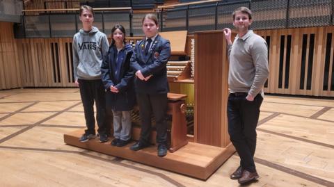 Three teenagers and a man are standing in front of an organ on a stage, looking at the camera.