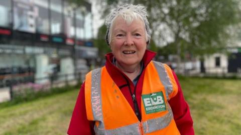 A woman in an orange high-vis jacket stands on a patch of grass with shops in the background.
