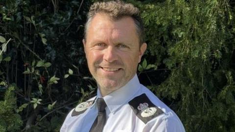 A man wearing a police uniform stands outside. Bushes can be seen behind him. He has short dark hair and stubble on his chin and cheeks.