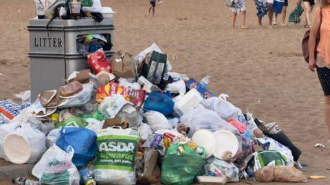 Silver waste bin with 'litter' written on it and litter overflowing on top and around it. It is on a sandy beach. There are people in the background.