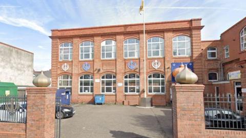 A rectangular brick building with large windows and colourful decorations on the wall. There is a car park with a flag pole in front of it and a gateway with two decorated brick posts