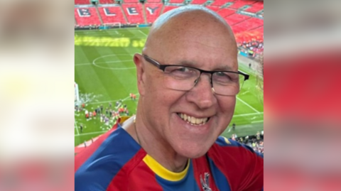 Rob Best. He is pictured standing in the stalls at Wembley Stadium. The football pitch can be seen in the distance behind him. Rob is wearing a football shirt and reading glasses. He is looking directly at the camera and smiling