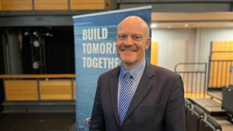 Deputy Gavin St Pier - A bald man, with a blue suit and a blue shirt, wearing a blue and white spotted tie. He is standing in front of a banner which says Build Tomorrow Together. 
