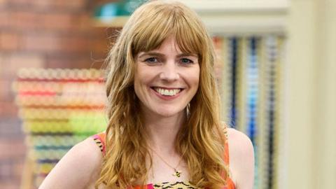 Close up of Adele, who has shoulder-length auburn hair stands in a brightly lit haberdashery area in the Sewing Bee studio