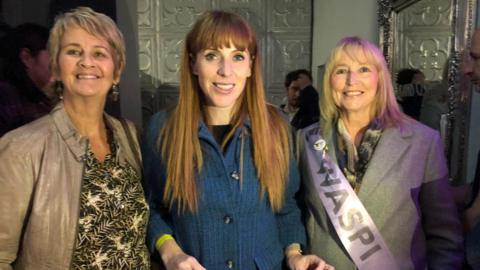 Three women smile at the camera, one woman on the right wears a WASPI sash