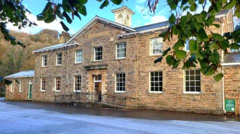 A historic brick building with two storeys. There are lots of sash windows on its frontage and a small ramp that leads up to the front door. There is also a large white clock tower on the roof. Outside the front of the building there is ice on a road surface, and in the background there is a forest of mature trees.