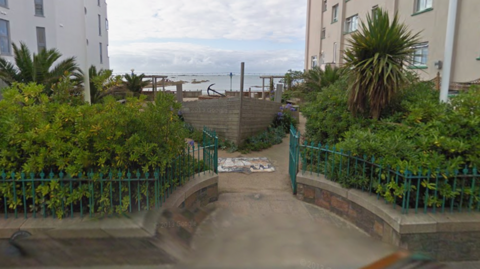 A picture of Allix's Shipyard. There is a number of plants and a wooden boat pictured in the yard. There is a body of water in the distance. There is two large buildings either side of the yard.